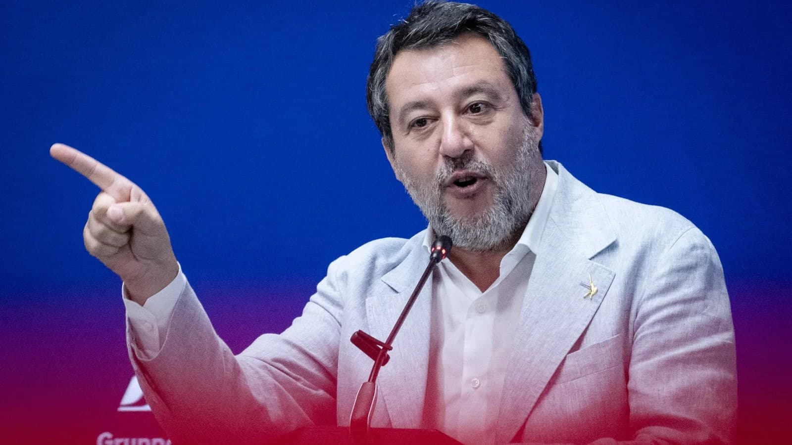 Matteo Salvini gesticola mentre parla da un podio su uno sfondo blu e rosso.